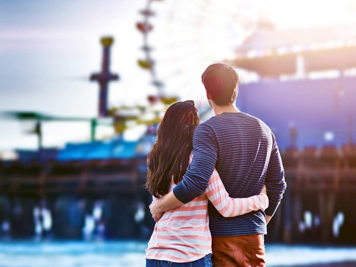 A couple stands by the beach, arms around each other, looking out at a ferris wheel and amusement park in the distance, enjoying the serene scenery.