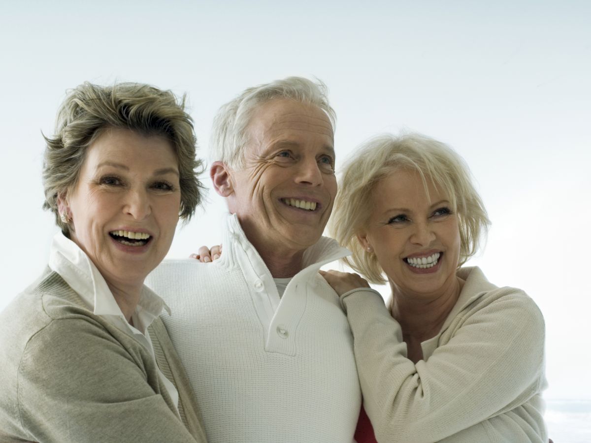 Three elderly people, two women and one man, are standing close together, smiling and embracing each other in a light background.