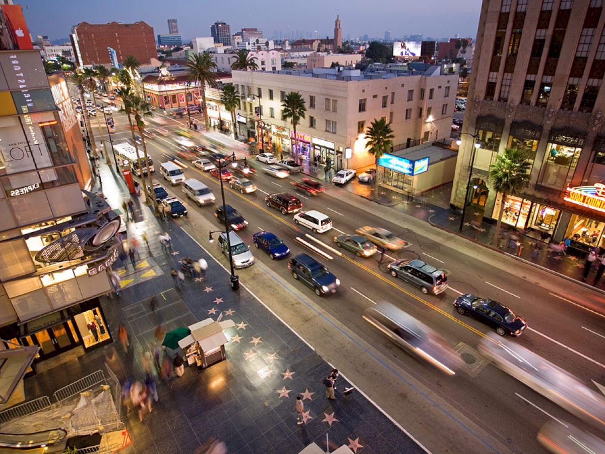 A busy street scene at dusk with cars, a sidewalk, and buildings lit by neon signs. The Walk of Fame stars are visible on the sidewalk.