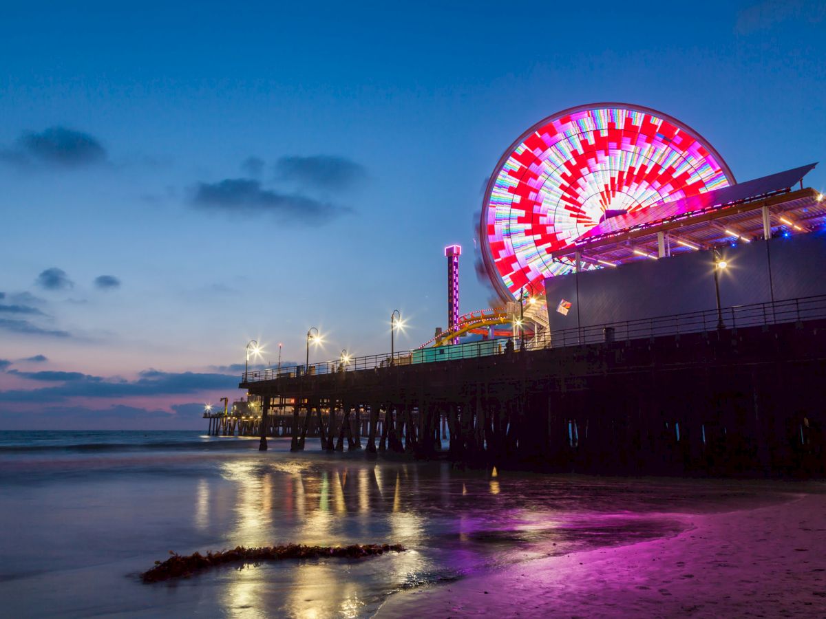 An illuminated Ferris wheel and amusement park on a pier during twilight, reflecting on the water and sandy shore.