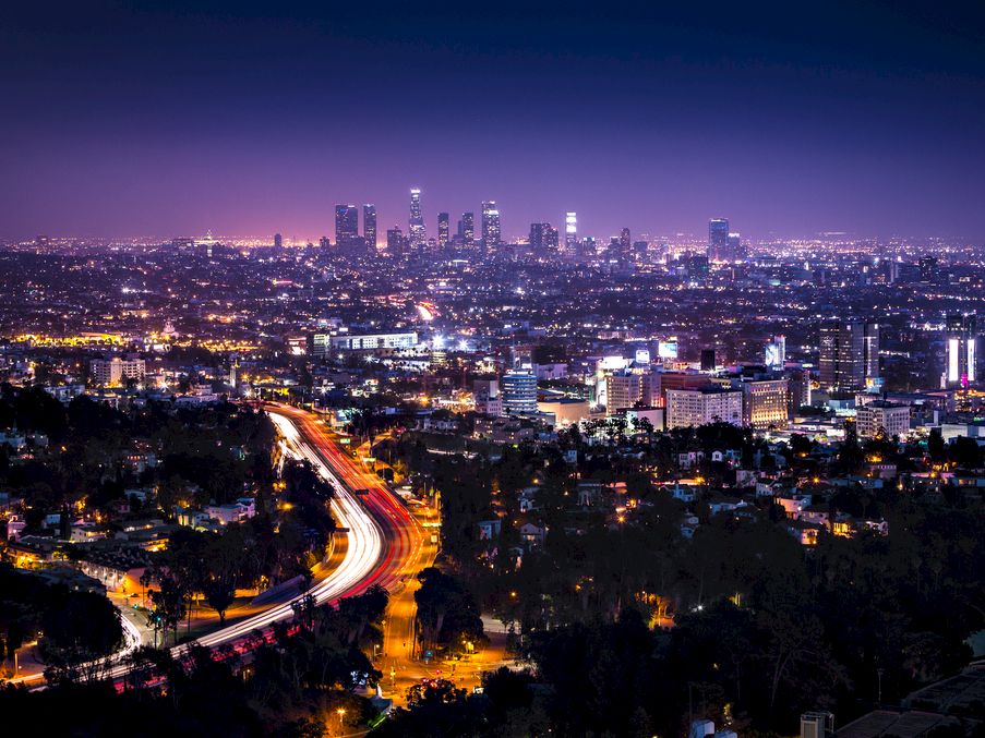 A vibrant cityscape at night, featuring illuminated buildings, a busy highway with streaking lights, all under a dark purple sky.