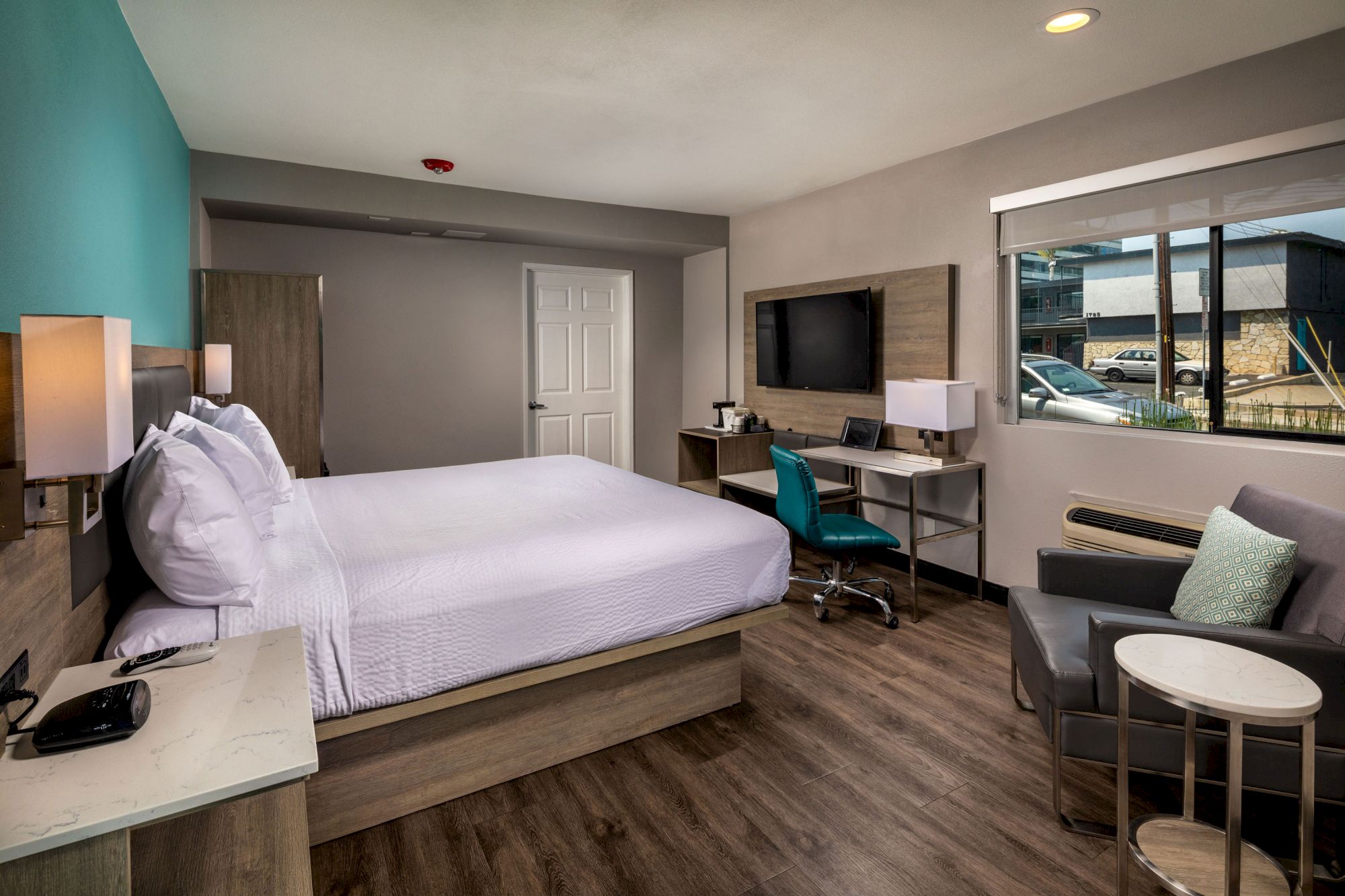 A modern hotel room with a queen-sized bed, desk, chair, TV, armchair, side table, lamp, and window view. The room has wooden flooring and teal accents.