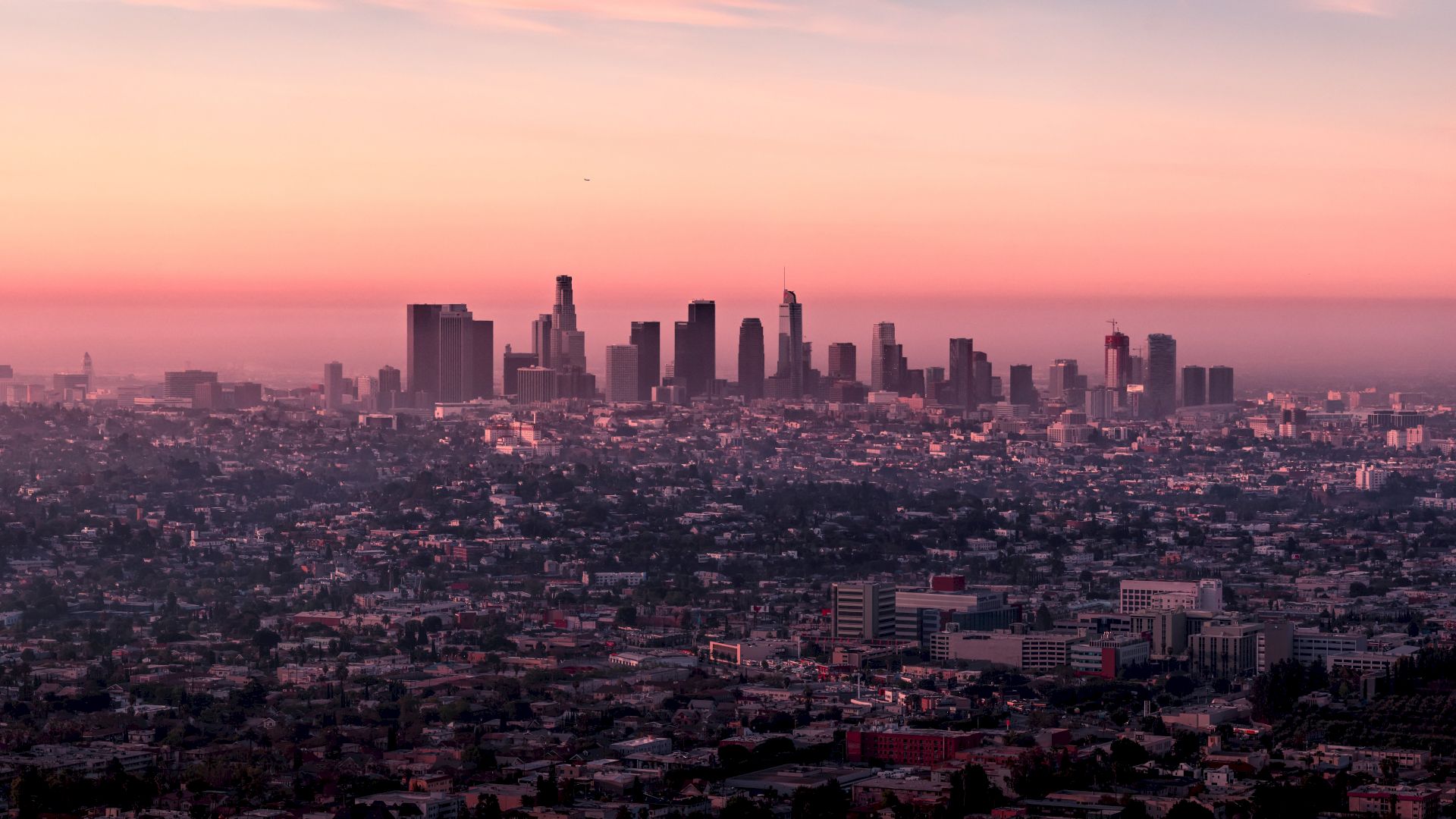 A panoramic view of a city skyline at sunset, with tall buildings outlined against a colorful sky and an expanse of cityscape below them.