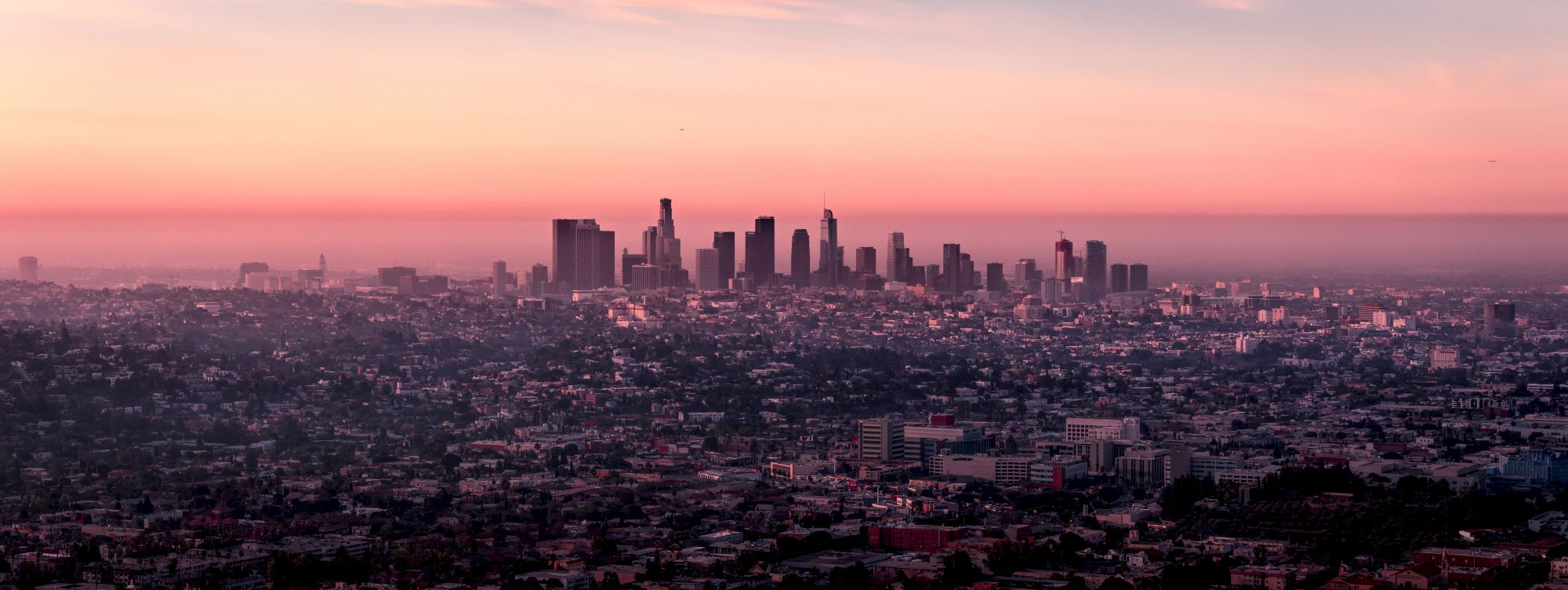 A panoramic view of a city skyline at sunset, with tall buildings outlined against a colorful sky and an expanse of cityscape below them.