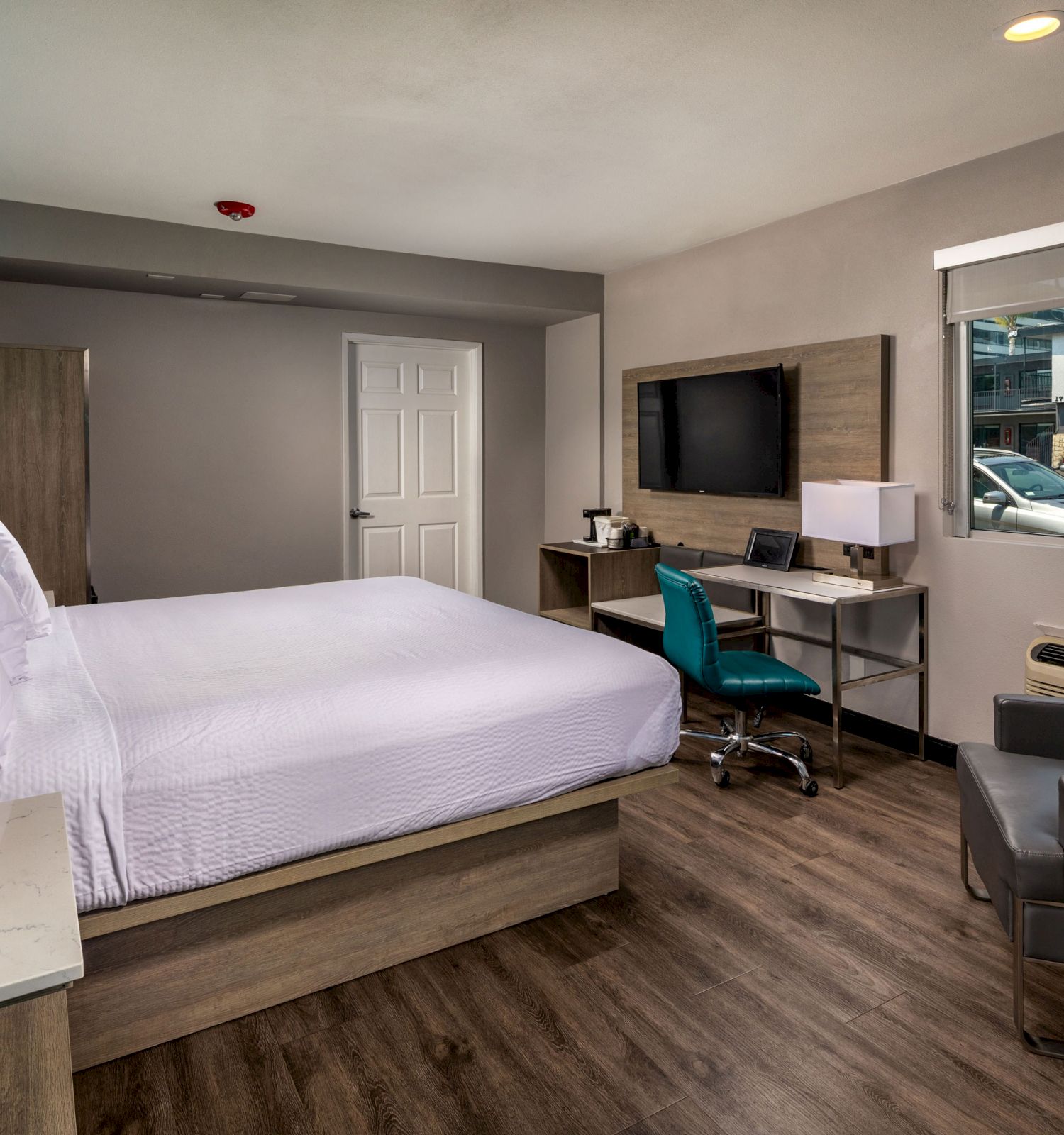 A modern hotel room with a double bed, desk, TV, armchair, coffee table, and a window showing a view outside, all on wooden flooring.