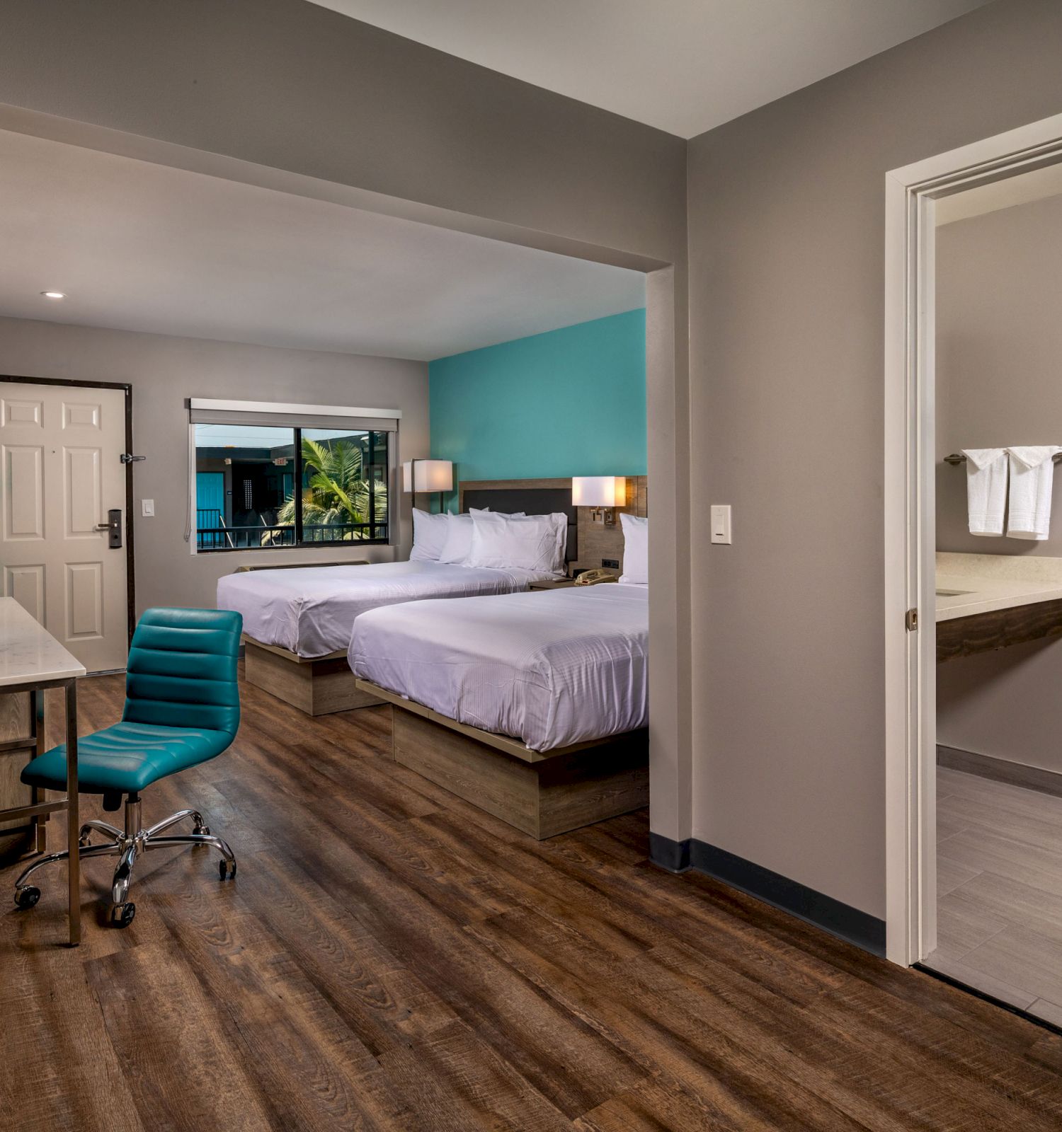 A modern hotel room with two beds, a work desk, a chair, and an adjoining bathroom. The room has hardwood floors and a teal accent wall.