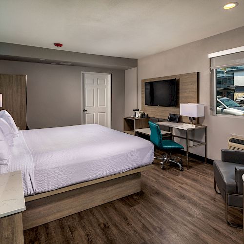 A modern hotel room with a large bed, desk, wall-mounted TV, window, and seating area with a chair and small table, featuring wood flooring.