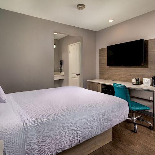 This image shows a modern hotel room with a bed, a wall-mounted TV, a desk with a chair, a coffee maker, and a view of the bathroom door.