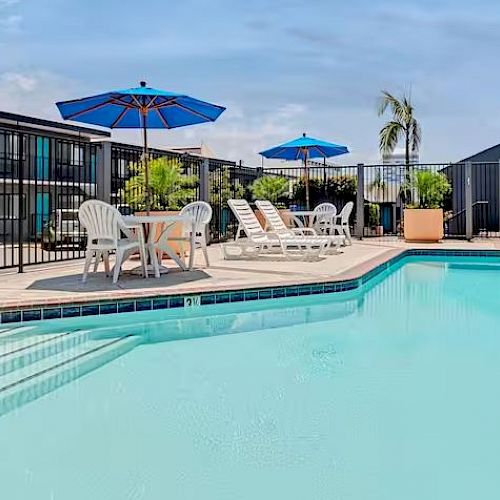 A swimming pool scene with lounge chairs, tables, umbrellas, and plants; enclosed by a black fence with a building in the background under a blue sky.