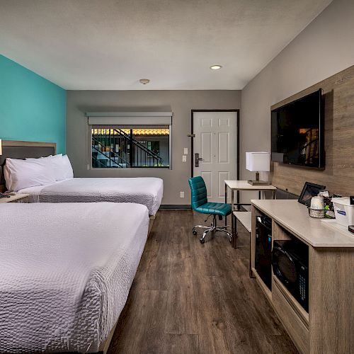 A modern hotel room with two beds, turquoise accent wall, TV, desk, chair, lamp, coffee maker, and bedside tables with lamps ends the sentence.