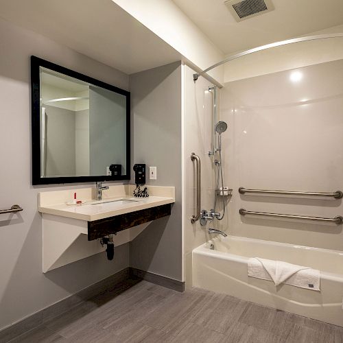 The image shows a clean, modern bathroom with a sink, a mirror, a bathtub with grab bars, and a neatly folded white towel on the tub.