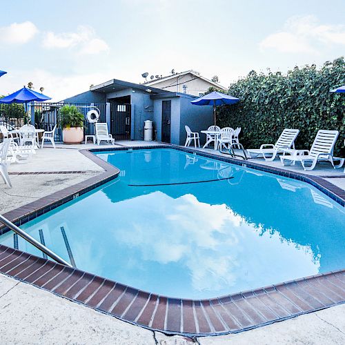 The image shows an outdoor swimming pool area with lounge chairs, tables, and umbrellas, all surrounded by greenery and a small building in the background.