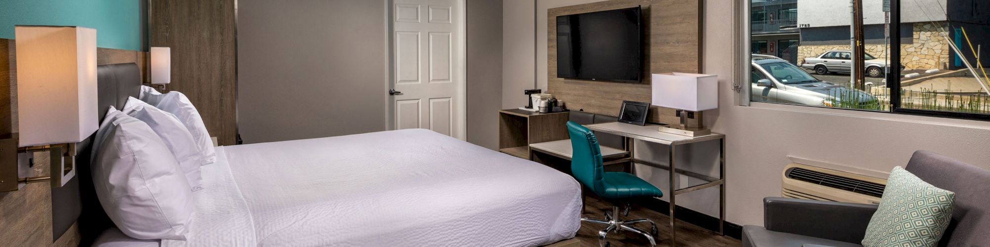 A modern hotel room with a large bed, desk, chair, TV, window, and sitting area with a table and armchair, featuring wood flooring.