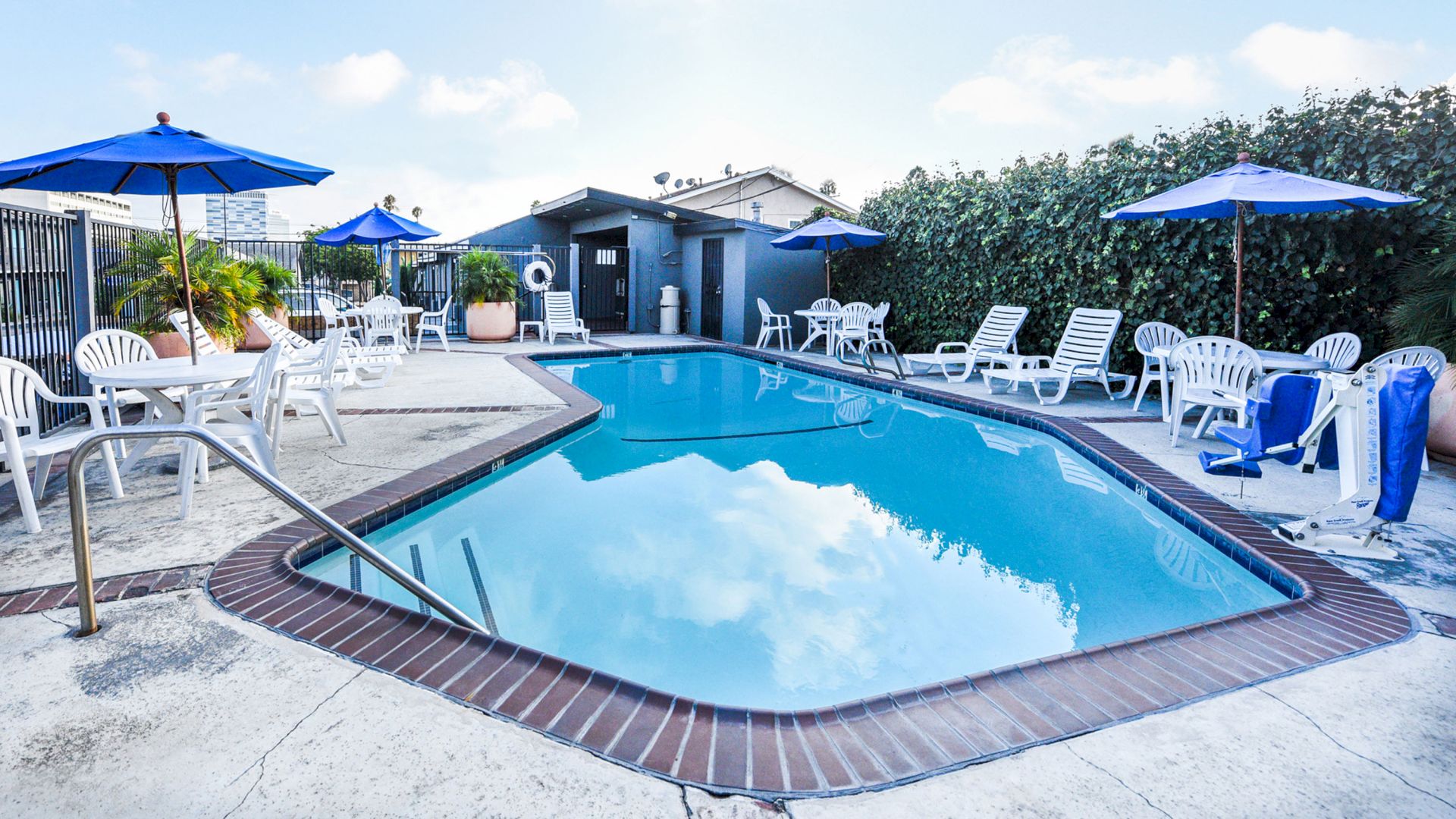 This image shows a swimming pool area with loungers, tables, and chairs, some with blue umbrellas. A small building is in the background.