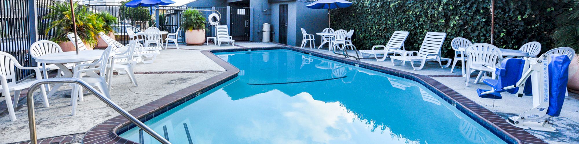 This image shows a swimming pool area with loungers, tables, and chairs, some with blue umbrellas. A small building is in the background.