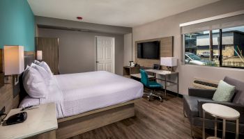 A modern hotel room has a bed, desk, chair, TV, and window; decor includes teal and neutral tones with wood flooring and contemporary furniture.