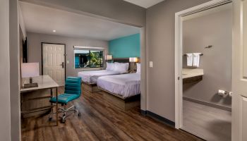 The image shows a hotel room featuring two beds, a desk with a chair, a lamp, and an adjacent bathroom with a sink and towels. The floor is wood-finished.