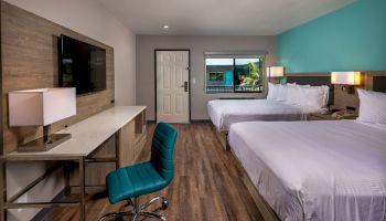 A hotel room features two beds, a desk with a lamp, a TV, a teal chair, and a window next to the entrance door.