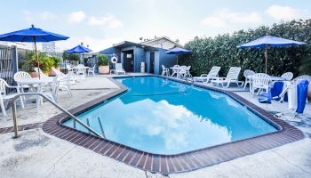 A swimming pool area with white lounge chairs, blue umbrellas, tables, and a small building in the background surrounded by a fence and trees.