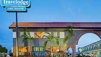 The image shows a Travelodge hotel with a sign that reads "Travelodge LAX South". The building is two stories high, with palm trees in front.