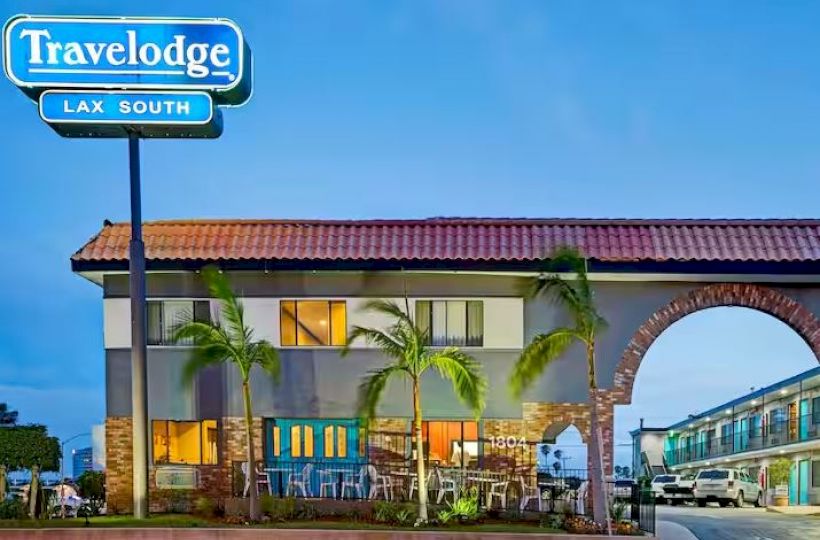 The image shows a Travelodge hotel with a sign that reads "Travelodge LAX South". The building is two stories high, with palm trees in front.